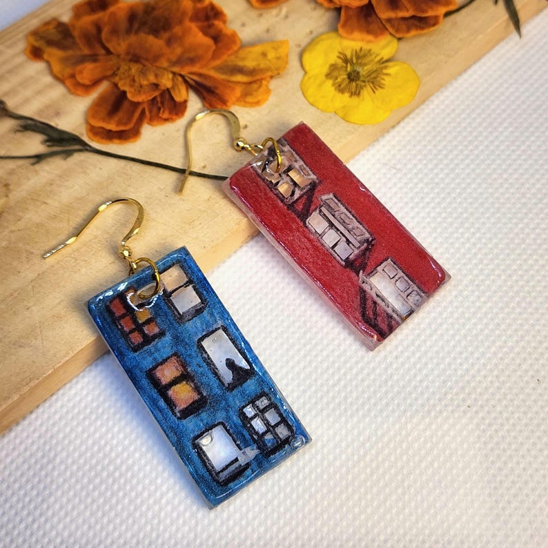 May include: A pair of rectangular drop earrings featuring hand-painted illustrations of building facades. One earring is blue with multiple window details, and the other is red with similar window patterns. Both earrings have gold-tone metal hooks.