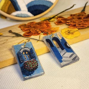 Pu&ograve; includere: Un paio di orecchini rettangolari in resina con scene di architettura mediterranea dipinte a mano. Un orecchino raffigura una finestra blu con fioriera, l'altro un portale ad arco blu con gradini in pietra e ganci in metallo dorato.