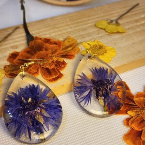 Può includere: Un paio di orecchini in resina a forma di goccia con petali di fiordaliso blu intenso incastonati. Ogni orecchino è appeso a un gancio in metallo dorato. La resina trasparente preserva la consistenza naturale e il colore vivido.