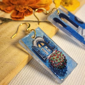 Pu&ograve; includere: Un paio di orecchini rettangolari in resina con una scena dipinta a mano di una porta ad arco blu con finiture bianche e un cespuglio di fiori rossi. Gli orecchini sono sospesi a ganci in metallo dorato e hanno una finitura lucida.
