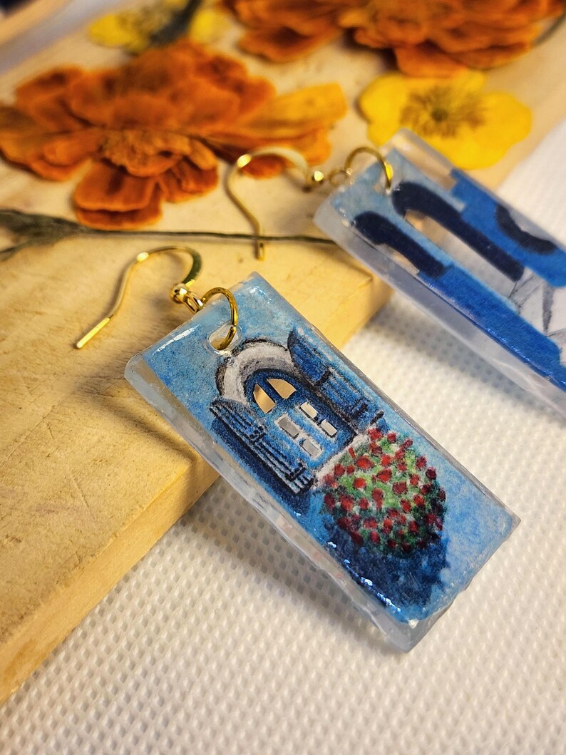 Pu&ograve; includere: Un paio di orecchini rettangolari in resina con una scena dipinta a mano di una porta ad arco blu con finiture bianche e un cespuglio di fiori rossi. Gli orecchini sono sospesi a ganci in metallo dorato e hanno una finitura lucida.