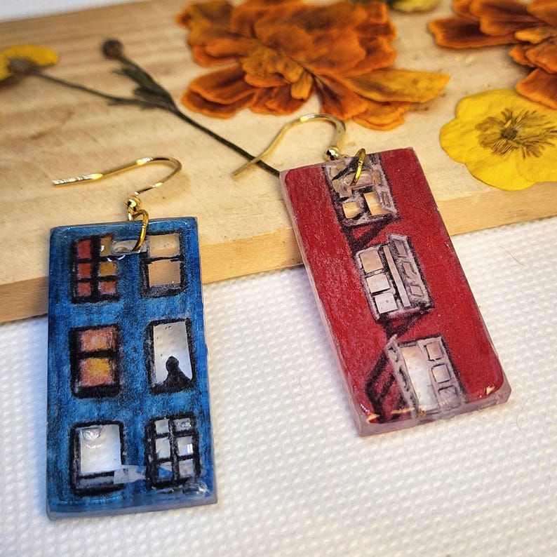 May include: A pair of rectangular drop earrings featuring hand-drawn illustrations of building facades. One earring is blue with multiple window panes, while the other is red with a similar window design. Both have gold-toned metal hooks.