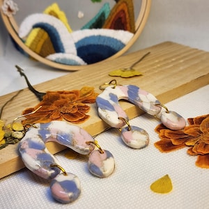 Può includere: Un paio di orecchini pendenti a forma di arco con un motivo marmorizzato nei toni del bianco, rosa polvere e blu pervinca con accenti dorati. Ogni orecchino è composto da un arco curvo con due piccoli ciondoli circolari fissati da anelli.