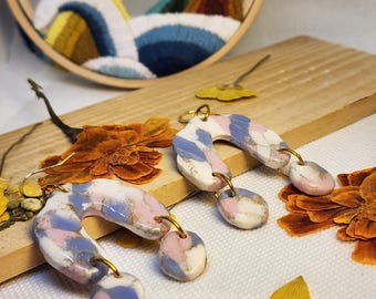 May include: A pair of arch-shaped dangle earrings featuring a marbled pattern in shades of white, dusty pink, and periwinkle blue with gold-toned accents. Each earring consists of a curved arch piece with two small circular charms attached by gold rings.