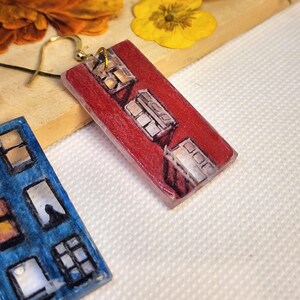 May include: A pair of rectangular drop earrings featuring hand-drawn illustrations of building facades. One earring is red with black-outlined windows, and the other is blue with black-outlined windows. Both have gold-tone metal earring hooks.