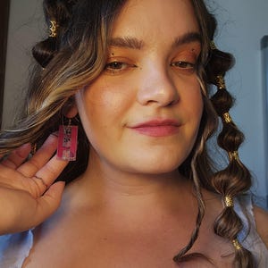 May include: A rectangular earring featuring a translucent pink acrylic base with a dark graphic print, suspended from a gold-toned metal hook. The earring displays a clear, glossy finish and a flat, geometric shape that hangs vertically from the earlobe.
