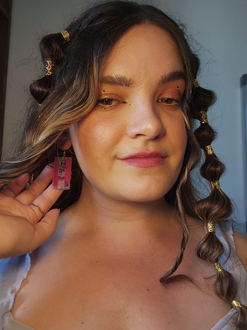 May include: A rectangular earring featuring a translucent pink acrylic base with a dark graphic print, suspended from a gold-toned metal hook. The earring displays a clear, glossy finish and a flat, geometric shape that hangs vertically from the earlobe.