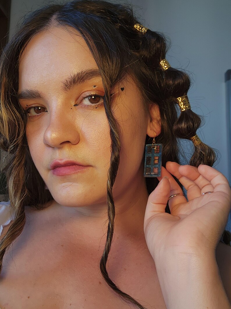 May include: A rectangular dangle earring featuring a blue frame with a grid of small, multi-colored square compartments resembling a makeup palette. The earring hangs from a gold-toned metal hook and displays a variety of warm-toned shades.
