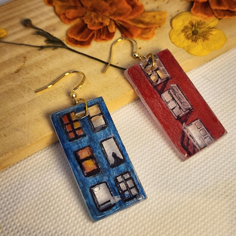 May include: A pair of rectangular drop earrings featuring gold-tone hooks. One earring is blue with a painted illustration of multiple windows, while the other is red with a similar painted window design. Both pieces have a smooth, glossy finish.