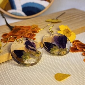Può includere: Un paio di orecchini rotondi in resina trasparente con petali di fiori viola incorporati e piccole macchie bianche. Ogni orecchino è fissato a un gancio in metallo dorato. Il design mostra elementi botanici naturali sospesi in un materiale liscio, circolare e trasparente, evidenziando il contrasto tra i petali viola scuro e la base in resina trasparente.