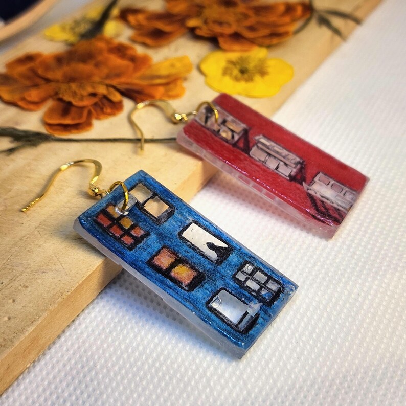 May include: A pair of rectangular drop earrings featuring hand-painted illustrations of building facades. One earring is blue with multiple window panes, while the other is red with a similar architectural design. Both have gold-tone metal hooks.