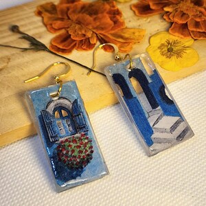 Pu&ograve; includere: Un paio di orecchini rettangolari in resina con ganci dorati. Un orecchino raffigura una finestra ad arco blu con fiori rossi, l'altro mostra colonne architettoniche blu e scale bianche, entrambi con una finitura lucida e liscia.