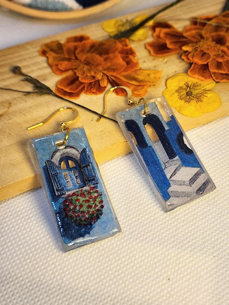 Pu&ograve; includere: Un paio di orecchini rettangolari in resina con ganci dorati. Un orecchino raffigura una finestra ad arco blu con fiori rossi, l'altro mostra colonne architettoniche blu e scale bianche, entrambi con una finitura lucida e liscia.