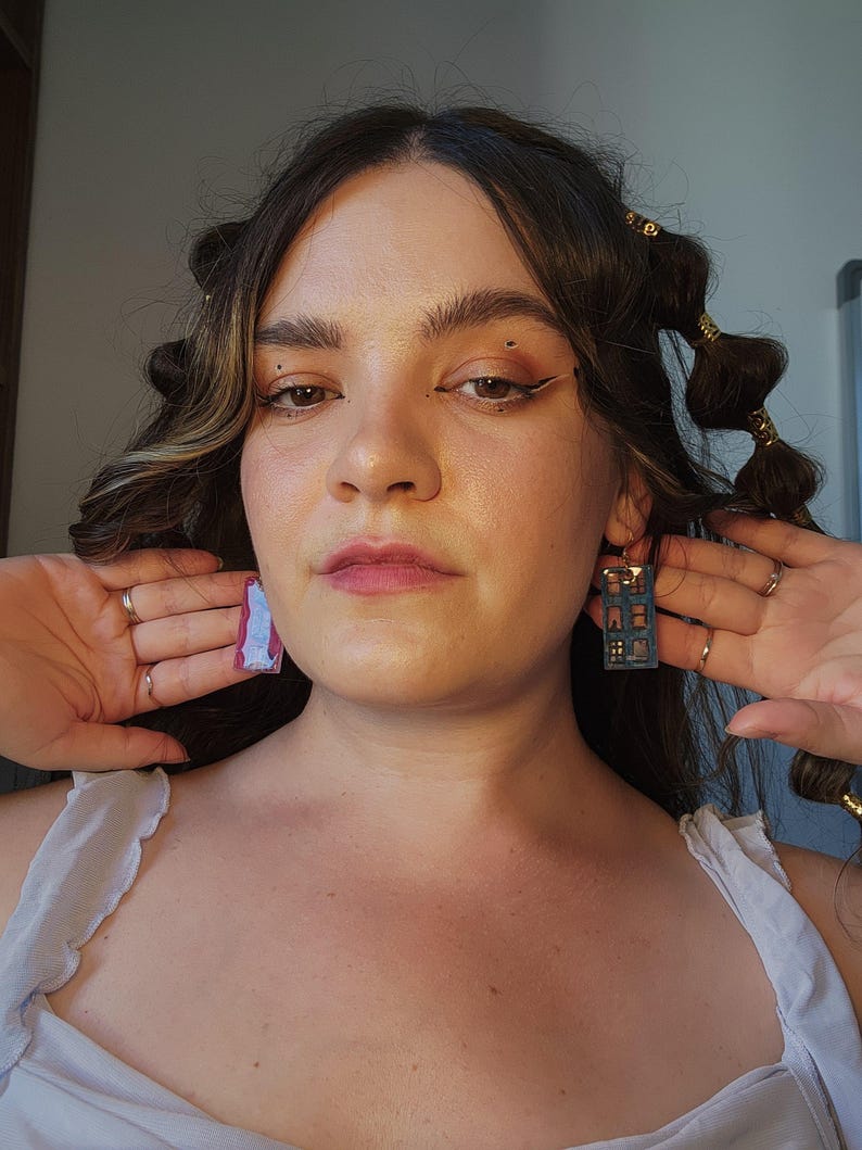 May include: A pair of mismatched rectangular earrings. One earring features a pink and white marbled pattern, while the other is a dark blue rectangular frame with a grid-like cutout design resembling a window or building facade.