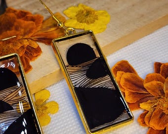 May include: A pair of rectangular drop earrings featuring a gold-toned metal frame. Each earring contains a clear resin inlay with a graphic design consisting of solid black circular shapes and fine, horizontal black lines on a white background.