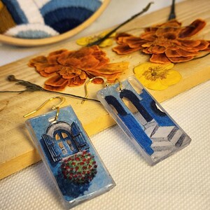 Pu&ograve; includere: Un paio di orecchini pendenti rettangolari in resina con ganci dorati. Un orecchino mostra una finestra ad arco blu con persiane aperte e fiori rossi, l'altro mostra un design di scala architettonica blu.