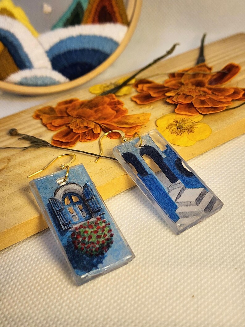 Pu&ograve; includere: Un paio di orecchini pendenti rettangolari in resina con ganci dorati. Un orecchino mostra una finestra ad arco blu con persiane aperte e fiori rossi, l'altro mostra un design di scala architettonica blu.