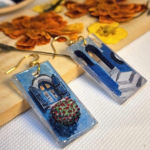 Pu&ograve; includere: Un paio di orecchini rettangolari con finitura in resina trasparente. Un orecchino raffigura una finestra blu con persiane e un cespuglio di fiori rossi, l'altro un arco architettonico blu con un pavimento a piastrelle geometriche.