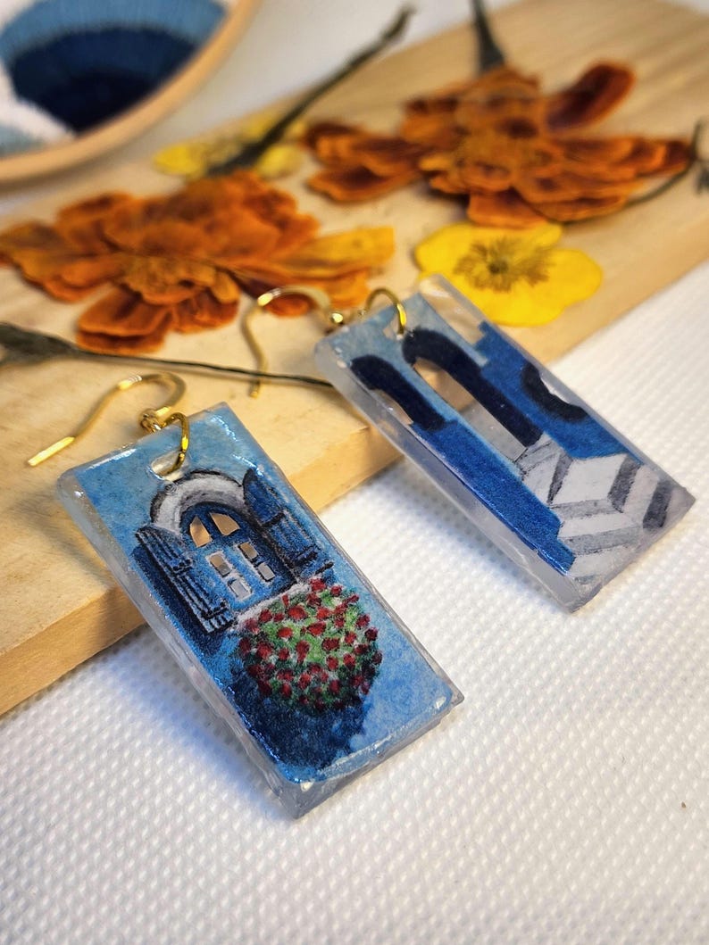 Pu&ograve; includere: Un paio di orecchini rettangolari con finitura in resina trasparente. Un orecchino raffigura una finestra blu con persiane e un cespuglio di fiori rossi, l'altro un arco architettonico blu con un pavimento a piastrelle geometriche.