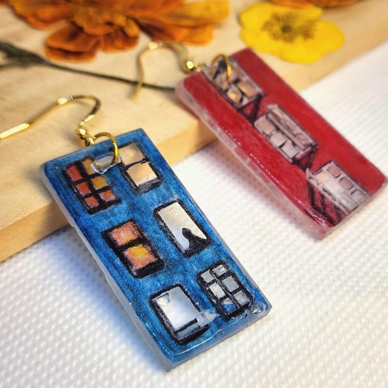 May include: A pair of rectangular drop earrings featuring hand-drawn illustrations of building facades. One earring is blue with multiple windows, and the other is red with similar window details. Both earrings have gold-toned metal hooks.