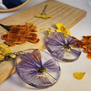 Può includere: Un paio di orecchini pendenti rotondi in resina trasparente con fiori pressati viola e piccole scaglie dorate incastonate. Ogni orecchino è fissato a un gancio in metallo dorato, mostrando un design botanico con petali naturali.