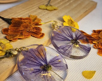 May include: A pair of round, clear resin drop earrings featuring embedded purple pressed flowers and small gold flakes. Each earring is attached to a gold-toned metal hook, showcasing a botanical design that captures the natural petals within.