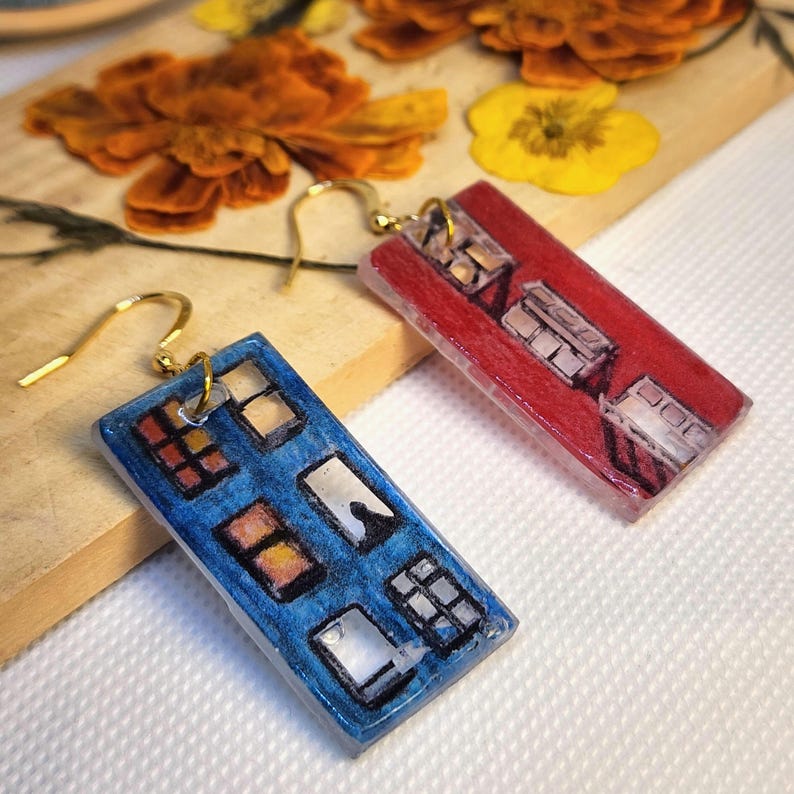 May include: A pair of rectangular drop earrings featuring a hand-painted design of building facades. One earring is blue with multiple window details, and the other is red with similar architectural window patterns. Both have gold-tone hooks.