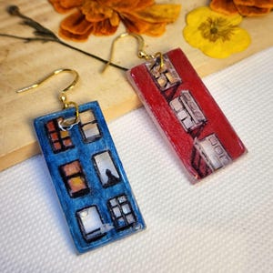 May include: A pair of rectangular drop earrings featuring a hand-painted building facade design. One earring is blue with multiple window panes, while the other is red with a similar window pattern. Both have gold-toned metal ear wire hooks.