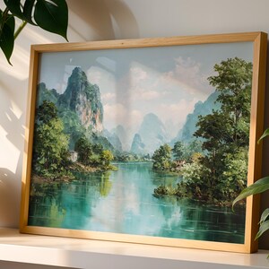 Può includere: Un dipinto di paesaggio incorniciato raffigura una serena scena fluviale con lussureggianti alberi verdi e imponenti montagne. L'opera d'arte presenta un cielo azzurro chiaro e una cornice in legno, creando un'estetica rilassante. Il dipinto è esposto su una mensola bianca.