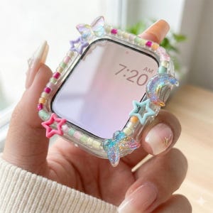 Puede incluir: Funda transparente para Apple Watch con un borde decorativo de cuentas de colores, estrellas, mariposas y una luna creciente. La esfera del reloj muestra la hora 7:20 AM. La funda se sostiene en una mano.