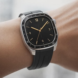 May include: A silver-toned wristwatch with a black face and black rubber band. The watch face has gold-colored hands and markers, with the number XII at the top. The watch is on a wrist.