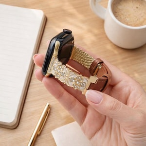 以下が含まれることがあります： 装飾的なバンドが付いたApple Watch。バンドは、ゴールドメッシュの中央部、クリアなラインストーンが付いた白いフローラルデザイン、茶色のレザーストラップが特徴です。時計の文字盤は、ゴールドフレームの黒色です。