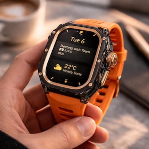 Könnte beinhalten: Eine orangefarbene Smartwatch mit schwarzem und goldenem Gehäuse. Das Display zeigt Datum, Uhrzeit und Wetter, einschließlich "22°C Mostly Sunny". Auf dem Bildschirm steht "Tue 6" und ein Meeting-Hinweis. Das Armband ist orange.