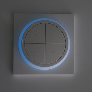 Könnte beinhalten: Ein quadratischer, weißer Lichtschalter mit vier kreisförmigen Knöpfen. Ein blaues Licht beleuchtet den kreisförmigen Rand um die Knöpfe. Der Schalter ist an einer grauen Wand montiert.