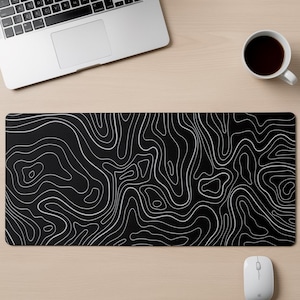 Può includere: Un tappetino da scrivania rettangolare con sfondo nero e un motivo a mappa topografica bianca composto da linee di contorno continue e fluide che coprono l'intera superficie, creando un design paesaggistico astratto e dettagliato.