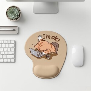 Puede incluir: Una alfombrilla de ratón ergonómica de color tostado con un dibujo de un gato atigrado naranja desplomado sobre un portátil con una pata levantada. El texto "I'm ok!" aparece en letras marrones sobre el gato. Incluye un reposamuñecas integrado.