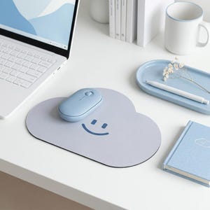 May include: A light blue cloud-shaped mouse pad featuring a dark blue smiling face graphic. The mouse pad is positioned on a white desk surface with a matching light blue wireless computer mouse resting on top of its smooth, rounded surface.