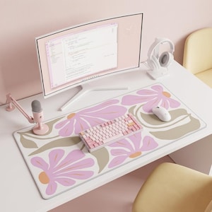 Op de afbeelding: Een grote rechthoekige bureauonderlegger met een herhalend patroon van gestileerde roze bloemen met oranje harten en beige bladeren op een witte achtergrond. De mat ligt op een wit bureau met een computermonitor, een roze microfoon, toetsenbord en muis.