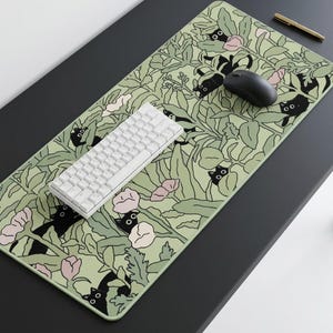 Pode incluir: Um tapete de secretária retangular com um fundo verde claro e um padrão repetitivo de gatos pretos escondidos entre folhagem verde estilizada e flores cor-de-rosa pálido. Um teclado mecânico branco e um rato de computador preto estão sobre o tapete.