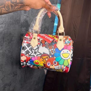 May include: A small barrel-shaped handbag featuring a vibrant, multi-colored graphic print with various cartoon characters, flowers, and star motifs. The bag has light beige top handles, one of which is wrapped in clear plastic, and a red tag.