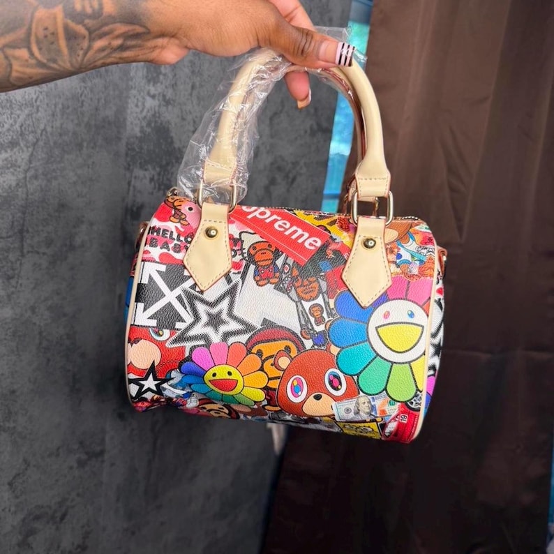 May include: A small barrel-shaped handbag featuring a vibrant, multi-colored graphic print with various cartoon characters, flowers, and star motifs. The bag has light beige top handles, one of which is wrapped in clear plastic, and a red tag.