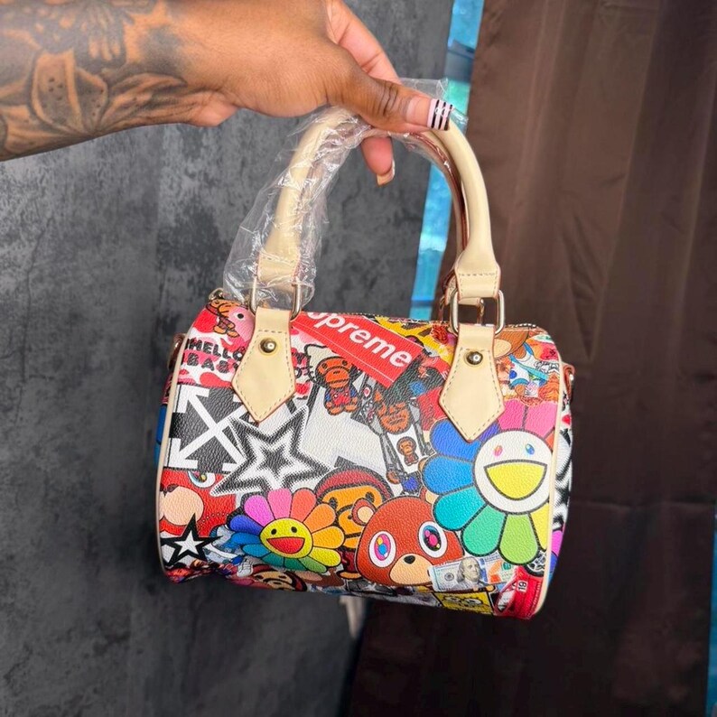 May include: A small barrel-shaped handbag featuring a vibrant, multi-colored graphic print with cartoon characters, flowers, stars, and a red Supreme logo tag. The bag has light beige leather handles, one of which is wrapped in protective plastic.