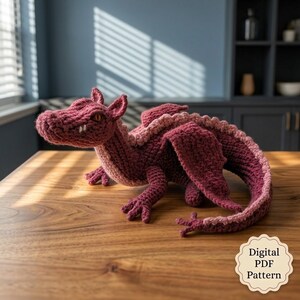 May include: A crocheted dragon toy in deep maroon with light pink accents along its back and tail. The creature features textured yarn, pointed ears, small white teeth, and orange eyes. A badge in the corner reads Digital PDF Pattern.