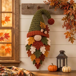 May include: A hanging crochet gnome decoration featuring a tall olive green hat with a rust-colored brim and a pom-pom tip. The gnome has a round beige nose and a long beard constructed from numerous crocheted autumn leaves in shades of orange, red, brown, and cream, accented with two small crocheted acorns.
