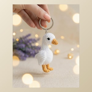 Puede incluir: Un llavero de ganso blanco tejido a ganchillo con pico y patas amarillas. El ganso tiene ojos negros y está sujeto a un llavero plateado. El fondo está borroso con iluminación suave y flores moradas.