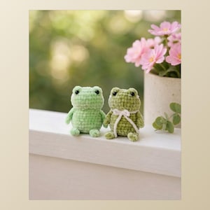 May include: Two small, handmade crochet frogs sit side-by-side. The left frog is light green, while the right frog is a darker olive green and features a small white yarn bow tied around its neck. Both have black bead eyes and round bodies.