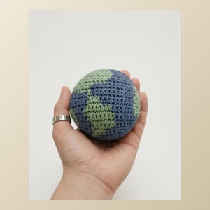 Puede incluir: Un globo terráqueo tejido a ganchillo en tonos azules y verdes, sostenido en una mano. Los continentes son verdes sobre un fondo azul. La esfera hecha a mano es una representación táctil de la Tierra.