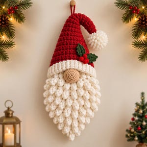 May include: A crocheted Christmas gnome wall hanging featuring a tall red hat with a white pom-pom, a beige round nose, and a long, textured white beard. The hat is decorated with two green crocheted holly leaves and three small red berries.