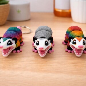 Könnte beinhalten: Drei farbenfrohe Opossum-Figuren mit offenem Mund, die einen regenbogenfarbenen Rücken, einen grauen Rücken und einen regenbogenfarbenen Rücken zeigen. Die Opossums haben rosa Pfoten und Nasen, schwarze Ohren und weiße Gesichter. Eines hat einen Schlüsselanhänger.