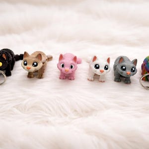 May include: Six cat figurines in a range of colours: black, brown, pink, white, grey, and rainbow. Each cat holds a silver ring in its mouth. The figurines are displayed against a white, fluffy background.